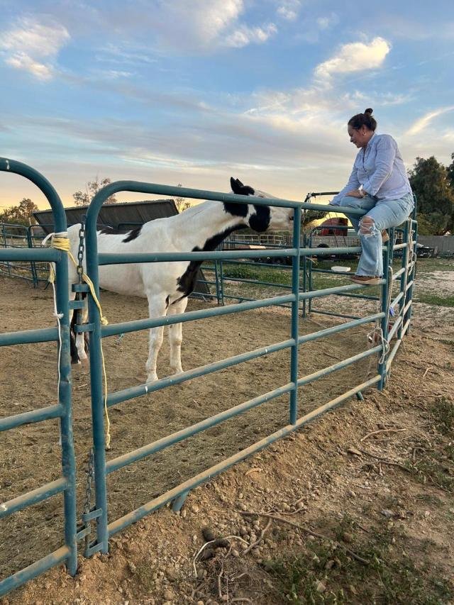 Alma sentada en la reja de un corral al atardecer, mirando a un caballo blanco y negro que se acerca a ella